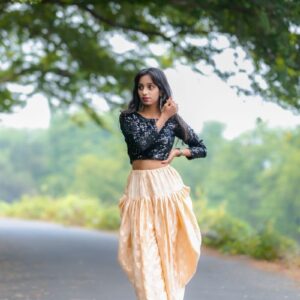 A beautiful woman in stylish attire stands confidently on a scenic road, surrounded by lush greenery.